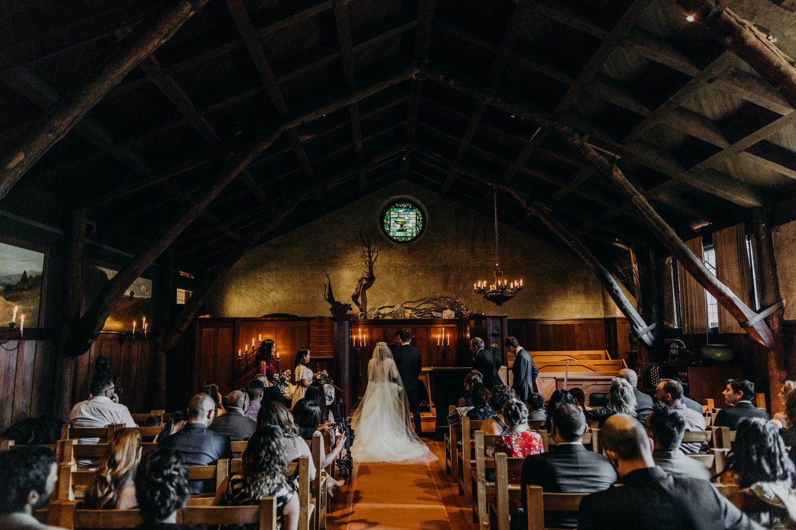 Inside the church during the ceremony: a high dark timber roof, guests in wooden pews on either side, and the bride in her veil at the far end of the aisle.