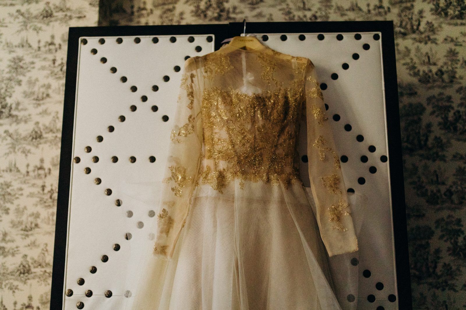 The wedding dress hanging on a wooden hanger against a white studded panel and grey toile wallpaper: a sheer champagne and gold lace bodice with long sleeves and a soft tulle skirt.