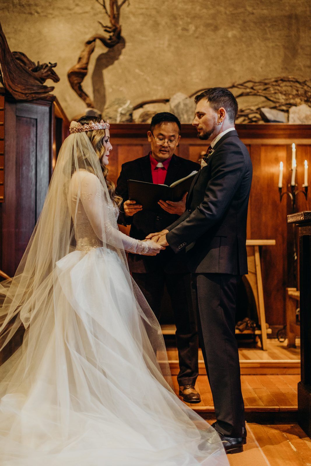 The bride in her long veil and the groom hold hands facing the officiant, who reads from a book, in front of a wall of stone and driftwood.