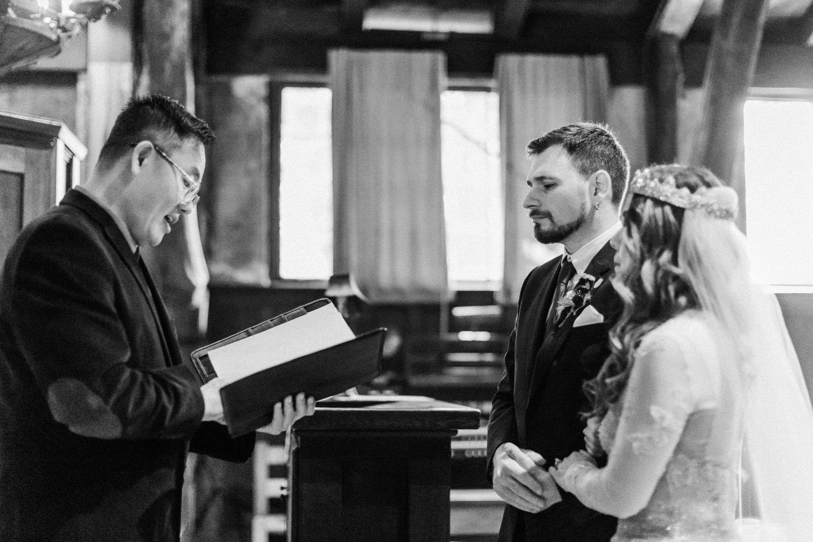 The officiant, smiling, reads from an open book at a wooden lectern while the groom and the bride in her veil stand facing him, in black and white.