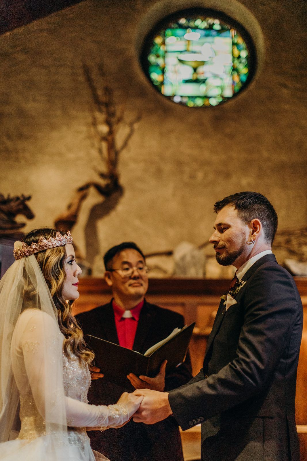 Rev. Junchol Lee smiling in his black robe and red stole as he joins hands with the couple at the altar, the round stained glass window and driftwood on the wall above them.