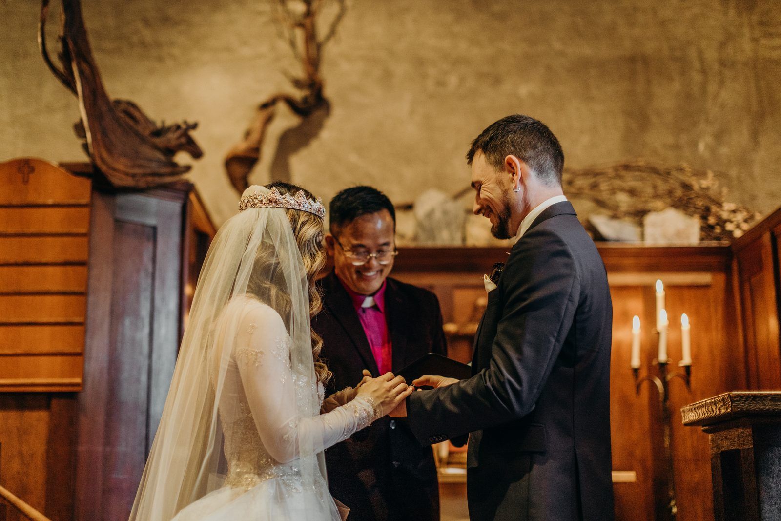 The bride places a ring on the groom's finger while the officiant looks on smiling, candles glowing on the wood panelled wall behind.
