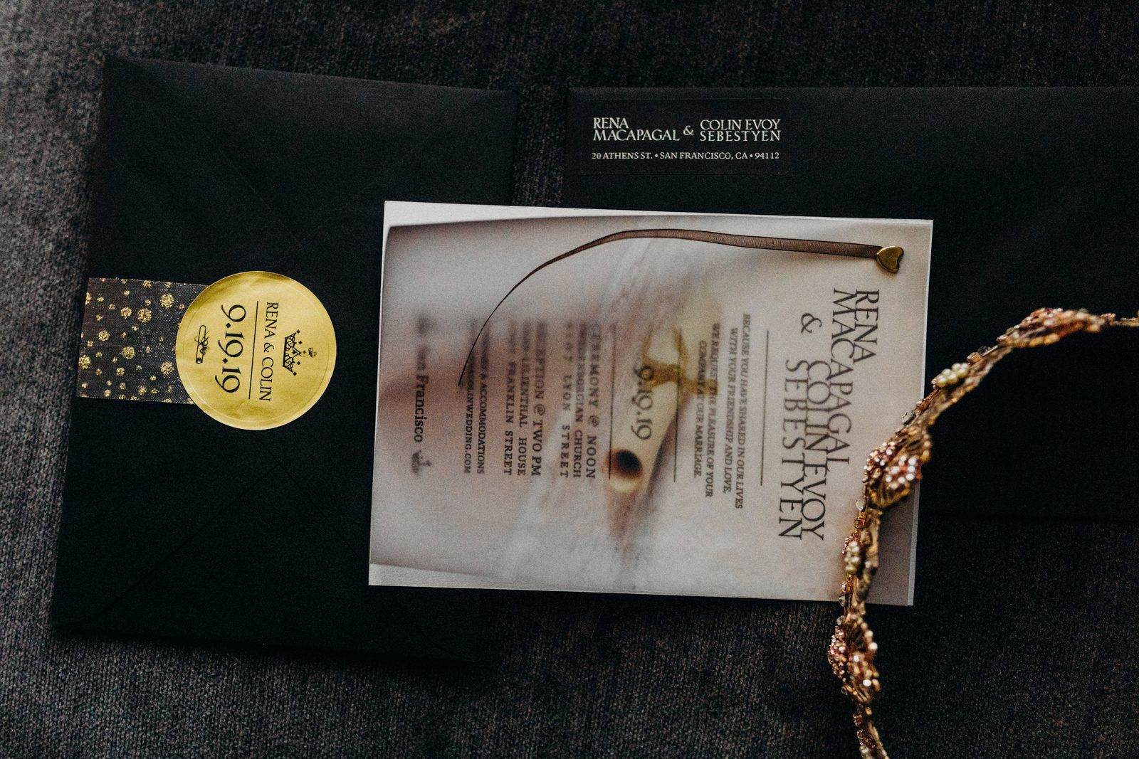 The invitation: a translucent vellum card with a brown ribbon and a small gold heart, lying on a black envelope sealed with a round gold sticker reading Rena & Colin 9.19.19, with the bride's gold crown at the edge of the frame.
