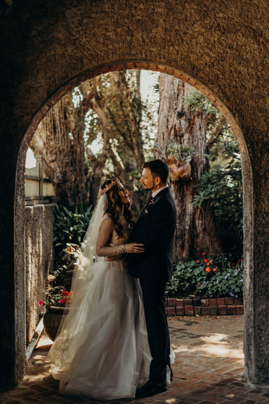 Rena and Colin standing close in a stone archway, framed by garden trees, Rena in her veil and crown looking up at Colin.