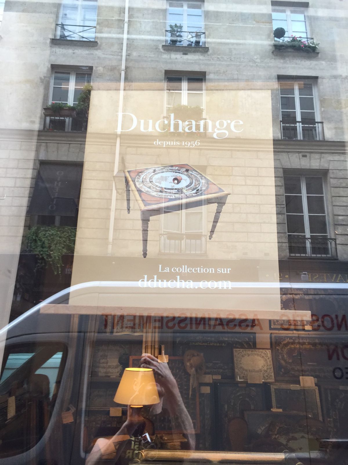 A shop window on a Paris street with the name Duchange and a drawing of a table on it, the street reflected in the glass.