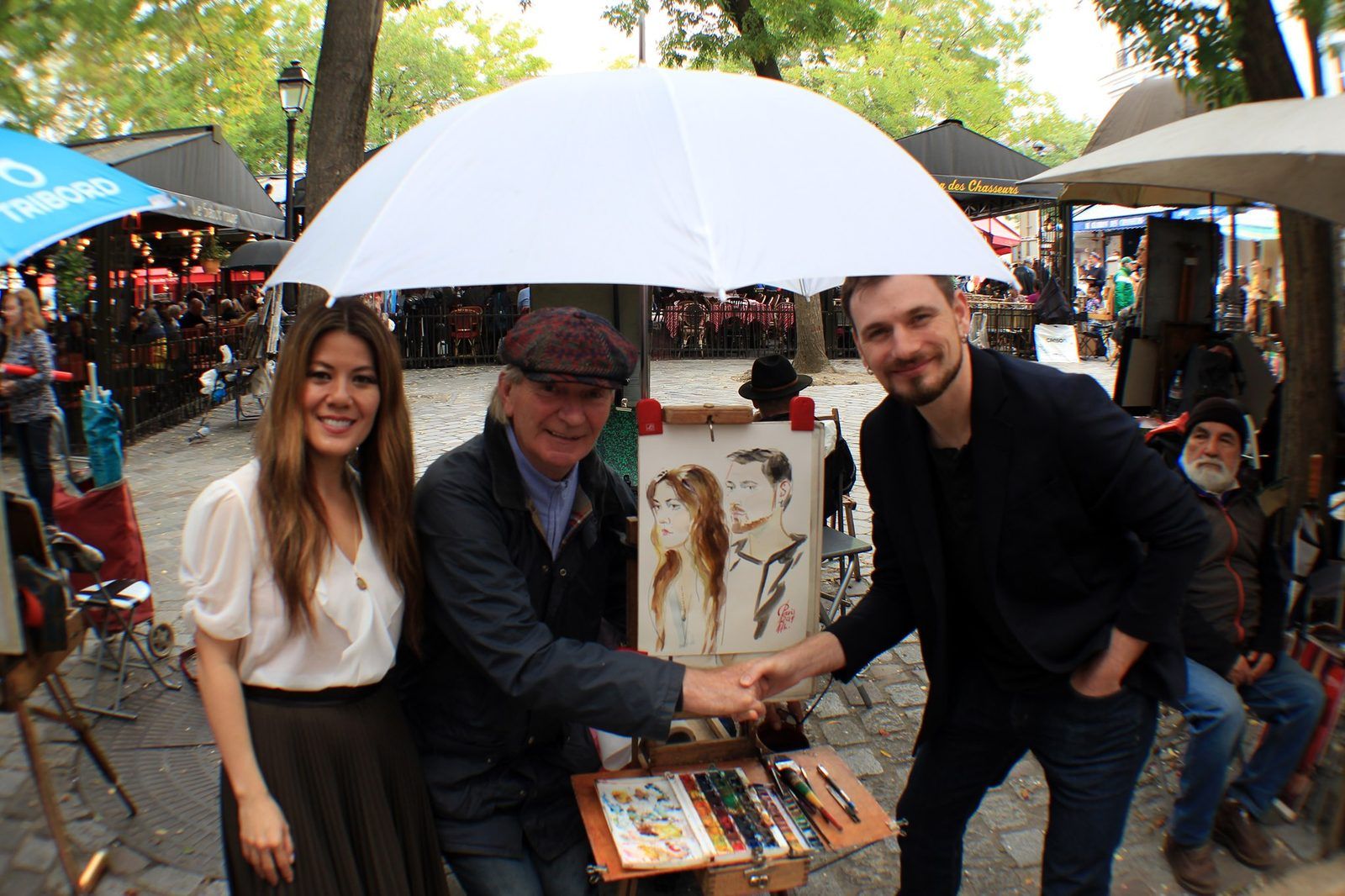 Rena and Colin standing with a street portrait artist under a white umbrella in Montmartre, shaking his hand, his watercolour portrait of the two of them on the easel between them.