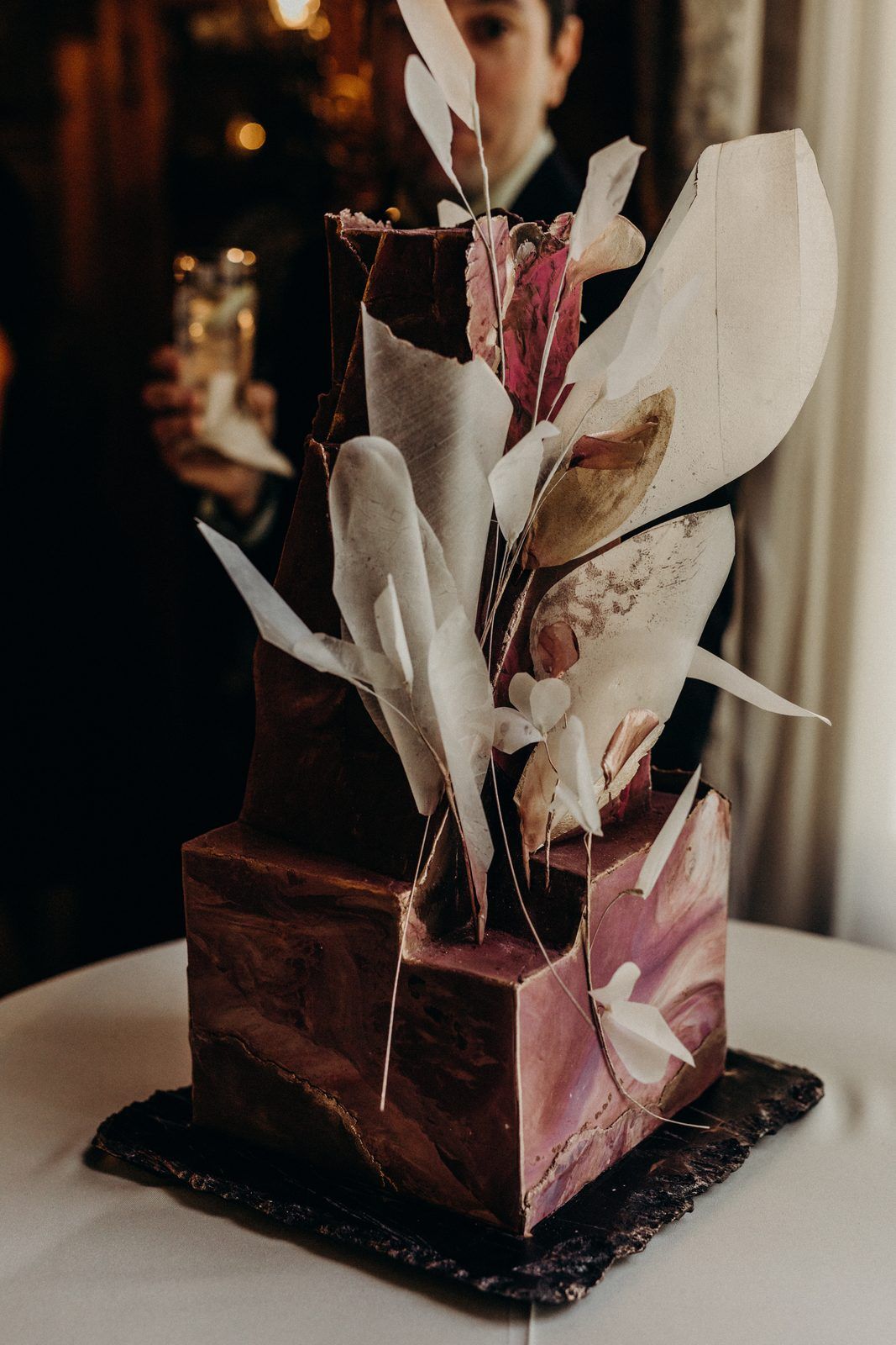 The finished cake on a slate slab: two square tiers marbled in plum and deep brown, with pale sugar leaves and fine stems rising out of the top.
