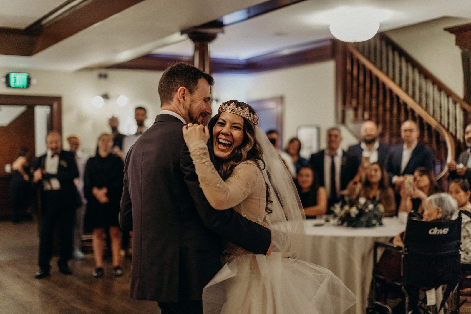 The couple dancing in the ballroom, the bride laughing with her head against the groom's shoulder while guests at round tables look on.