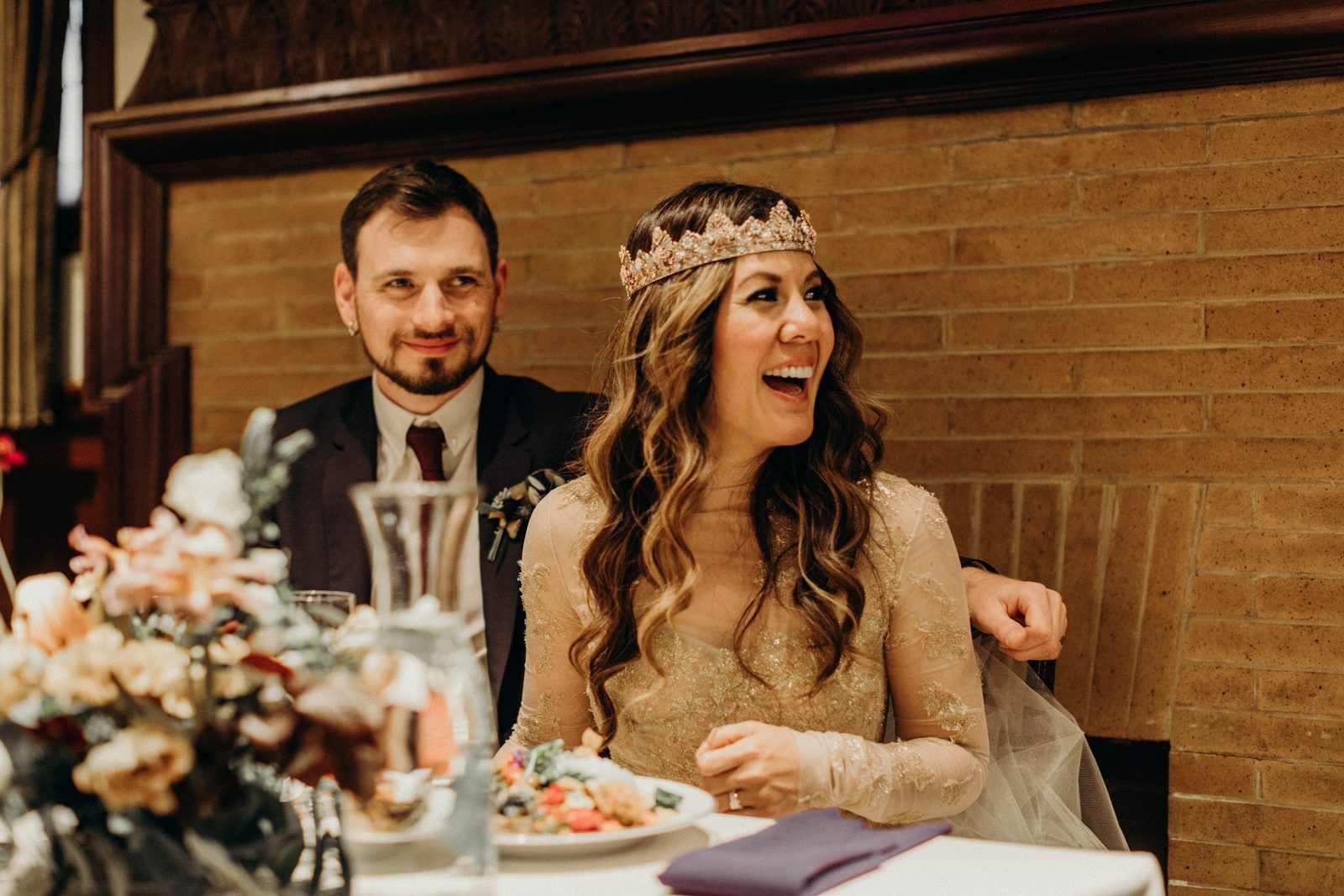 The couple at their table against a brick wall, the bride laughing in her crown and gold lace dress, the groom smiling beside her.