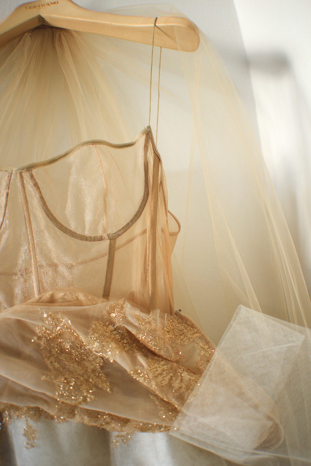 The champagne gold and nude lace bodice of the dress hanging on a wooden hanger, with sheer tulle over it.